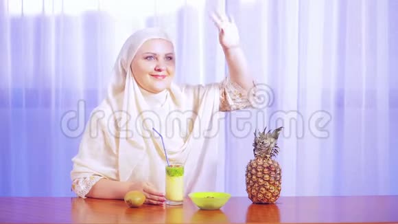 一位年轻的穆斯林妇女戴着一条浅色围巾从一根稻草上喝下一杯水果冰沙并向某人挥手致意视频的预览图