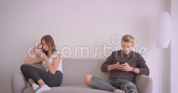 一对年轻可爱的白种人夫妇用手机分别坐在室内沙发上的特写镜头视频的预览图