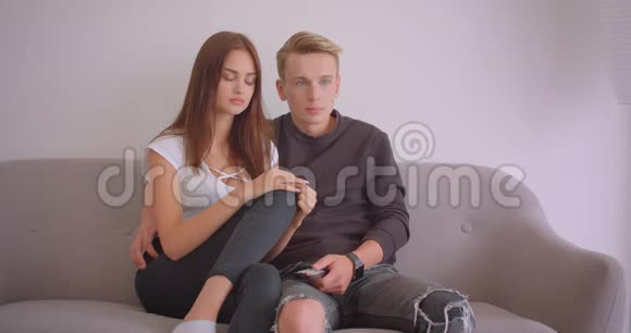一对年轻可爱的白种人夫妇在电视上看恐怖电影时被吓得坐在沙发上的特写镜头视频的预览图
