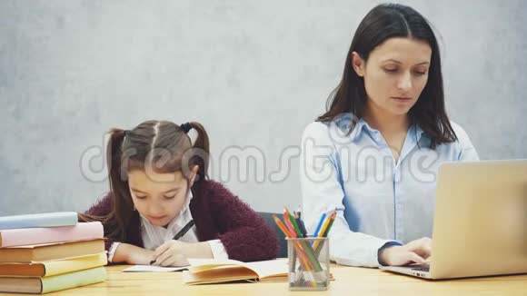 年轻的母亲坐在桌旁时在笔记本电脑上工作女儿在此期间写作业灰色背景视频的预览图