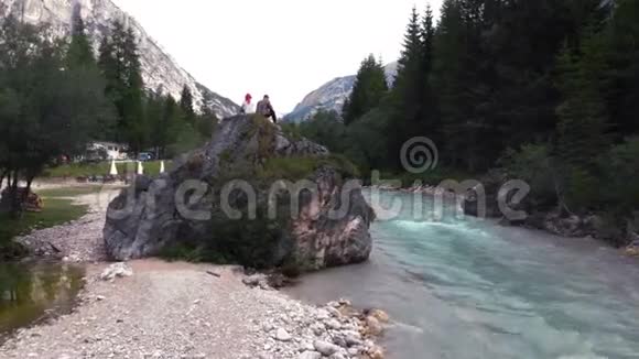 前景一对夫妇坐在一块大石头上一块巨石上在一条绿松石色的美丽河中多洛米特斯视频的预览图