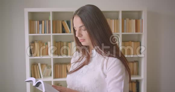年轻漂亮的白种人女学生正在看一本书看着相机在室内开心地笑着视频的预览图
