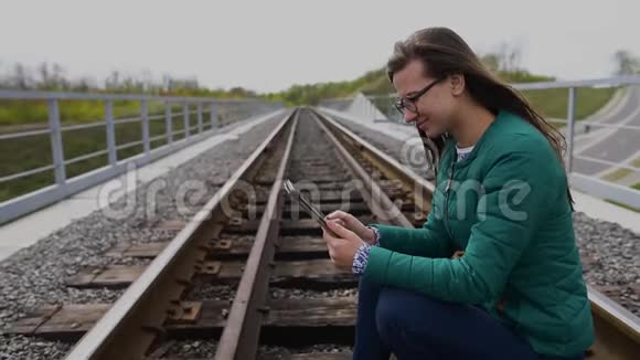 年轻的微笑女孩用平板电脑站在铁路旁戴着眼镜穿着绿色的衣服视频的预览图
