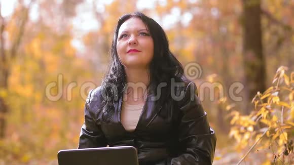 在一个阳光明媚的秋日一个年轻的黑发女人坐在公园里看着笔记本电脑屏幕微笑着视频的预览图