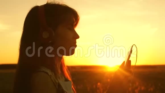 美丽的女人在美丽的夕阳下听音乐跳舞戴耳机的年轻女孩视频的预览图