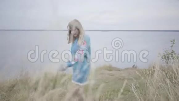 漂亮的金发女孩穿着漂亮的短蓝色夏装刺绣在草地上跳舞这条河在河上视频的预览图