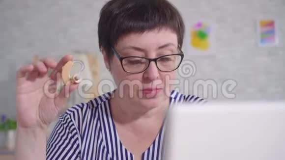 带眼镜的女人坐在客厅里的桌子旁手里拿着助听器用笔记本电脑合上视频的预览图