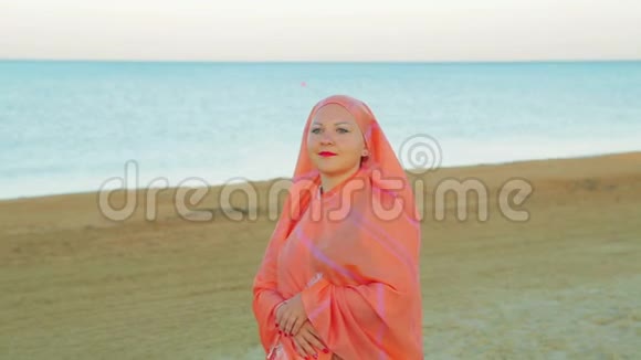 一个穿着橙色披肩的年轻穆斯林女子在沙滩上站在大海的背景下视频的预览图