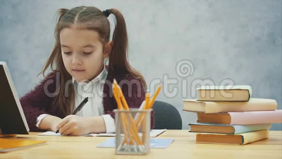 一个好的女学生坐在灰色背景的桌子旁在这期间他一边写作业一边看书视频的预览图