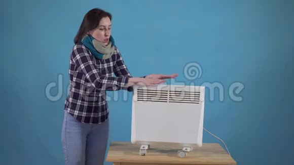 裹着围巾的冷冻女人然后用蓝色背景的电加热器加热视频的预览图