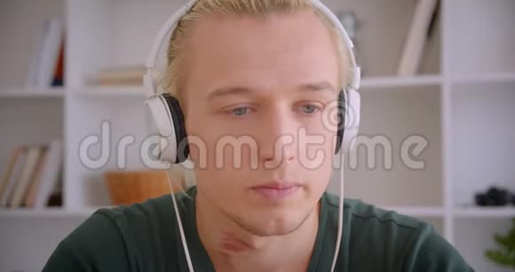 一位年轻迷人的金发时髦男士戴着耳机看着摄像机在室内听音乐视频的预览图