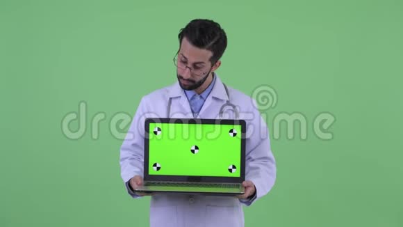 快乐的年轻留胡子的波斯人医生一边看着笔记本电脑一边思考视频的预览图