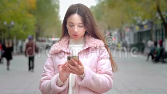一个年轻的女孩站在一条城市街道上拿着智能手机输入短信与朋友聊天视频的预览图