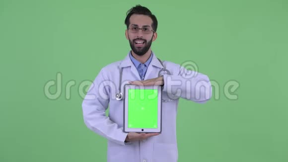 快乐的年轻人长着胡子的波斯人医生一边说话一边显示数字平板电脑视频的预览图