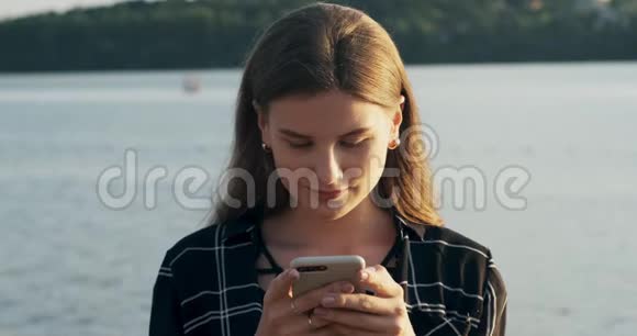 特写照片年轻迷人的女人微笑着在日落时看着海滩附近的智能手机自信的美丽视频的预览图