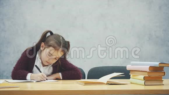 一个勤奋困倦的女儿写功课在这段时间里他坐在灰色背景的桌子旁掉下来了视频的预览图
