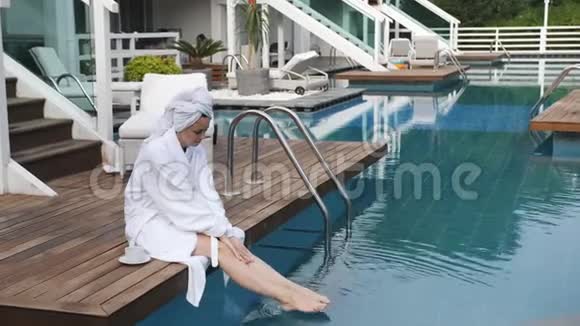 优雅的女人穿着白色浴袍坐在豪华别墅的游泳池旁的码头上喝着咖啡视频的预览图
