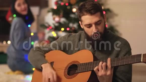 男人和女人坐在现代房间的圣诞树附近女孩挂玩具可爱有趣的男人弹吉他快乐夫妻视频的预览图