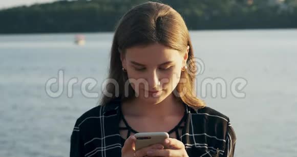 特写照片年轻迷人的女人微笑着在日落时看着海滩附近的智能手机自信的美丽视频的预览图