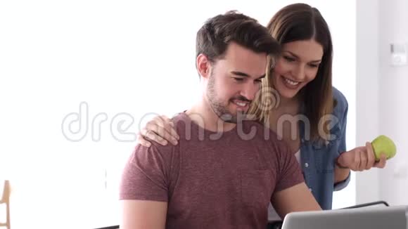 漂亮可爱的年轻夫妇用笔记本电脑在家里的厨房里聊天视频的预览图