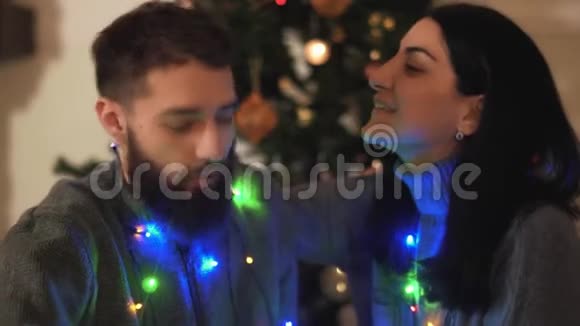 男人和女人坐在圣诞树附近脖子上挂着花环恋人们在说话微笑快乐视频的预览图
