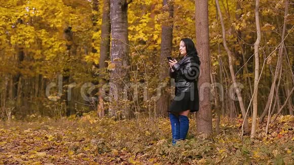 秋天公园里一个穿着蓝色靴子的年轻黑发女人站在一棵树旁在打电话视频的预览图