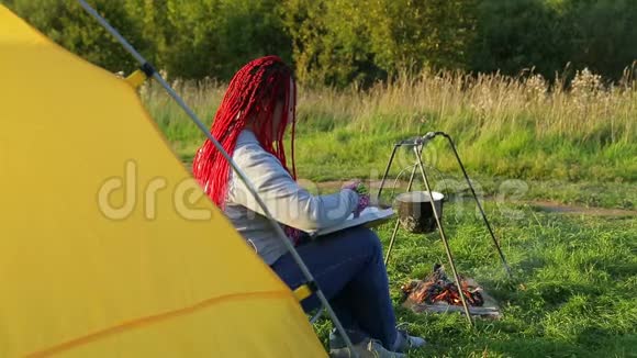 一个在篝火帐篷附近戴着非洲辫子的女人看着一个沸腾的锅视频的预览图