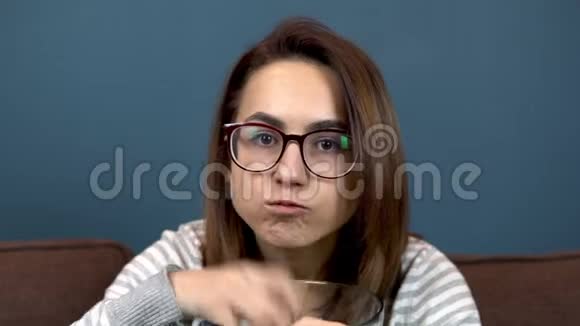 一个戴眼镜的年轻女人吃薯片看电视特写女孩积极吃芯片和开关频道视频的预览图
