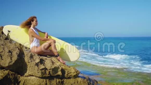 冲浪女孩坐在一个美丽的岩石海滩与木板强大的海浪冲击着岩石海岸女孩看着远处视频的预览图
