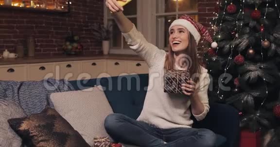美丽的笑容白人金发女孩戴着圣诞帽坐在沙发上圣诞家庭背景做自我视频的预览图