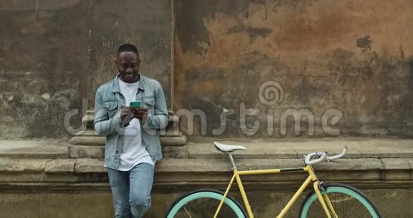 兴奋的美国黑人男子使用他的智能手机玩在线游戏同时站在附近的旧建筑墙边视频的预览图