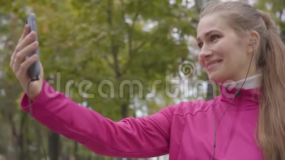 穿着粉红色运动服的白种人微笑的女人通过智能手机发送空气吻并开始跑步的肖像女性视频的预览图