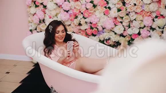 快乐美丽的女孩躺在白色的浴缸里手里拿着一部智能手机视频的预览图