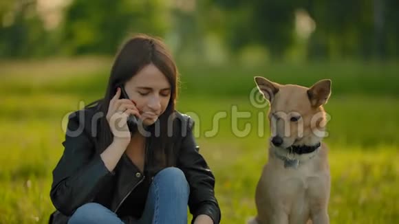 迷人的女孩在公园里和狗散步时在智能手机上说话视频的预览图