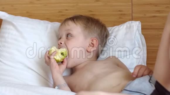一个金发小男孩躺在一张白色的床上吃着一个苹果视频的预览图