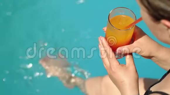 一个女人坐在游泳池边缘的特写镜头配上五颜六色的鸡尾酒视频的预览图
