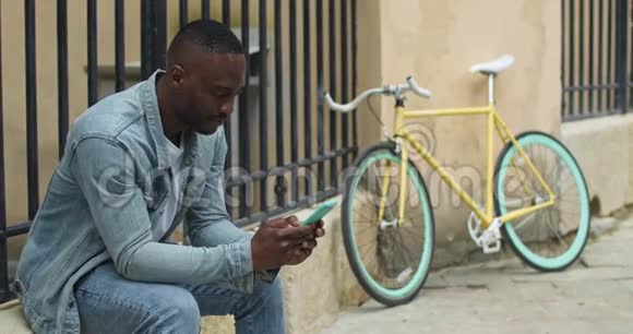 快乐的非裔美国人年轻人使用他的智能手机打字信息和朋友聊天而坐在时尚附近视频的预览图