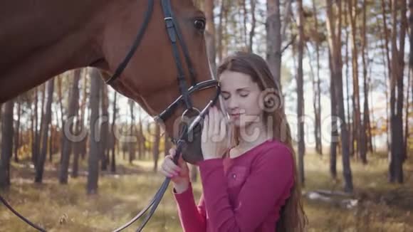 一个穿着粉红色衣服的年轻白种人女孩在秋天的森林里抚摸着马微笑着美丽的黑发视频的预览图