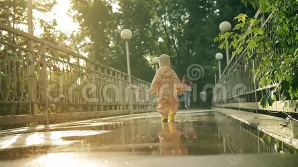 穿着橙色防水雨衣和橡胶雨靴的小女孩在水坑上向母亲走去慢镜头视频的预览图