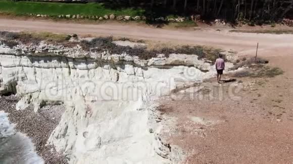 穿着短裤运动鞋和T恤的人沿着悬崖散步泡沫海浪撞击岩石海岸直到视频的预览图