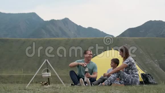 幸福的年轻家庭有一个孩子在山上的帐篷前休息喝热水瓶里的茶视频的预览图
