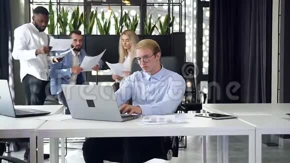 与他的同事在现代办公室的笔记本电脑上工作的一位戴着眼镜的英俊聪明的年轻商人亲密无间视频的预览图