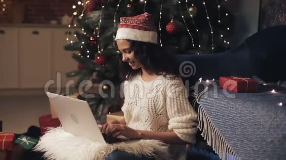 圣诞老人帽子里的漂亮年轻女人打字和使用笔记本电脑时坐在家门口的地板上视频的预览图