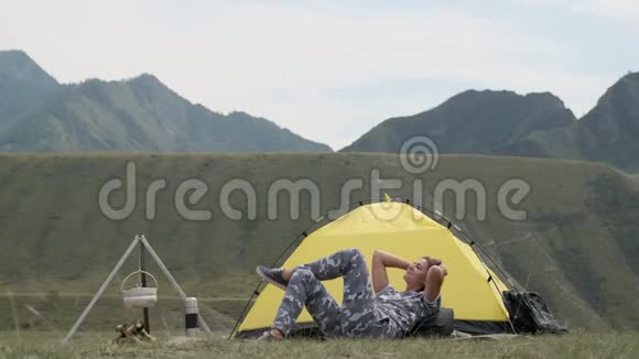 在晴朗的天气里身穿灰色衣服的女游客躺在帐篷前的山上放松视频的预览图