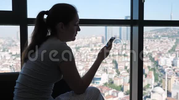 女人正在浏览智能手机坐在客厅里可以看到全景城市景色视频的预览图