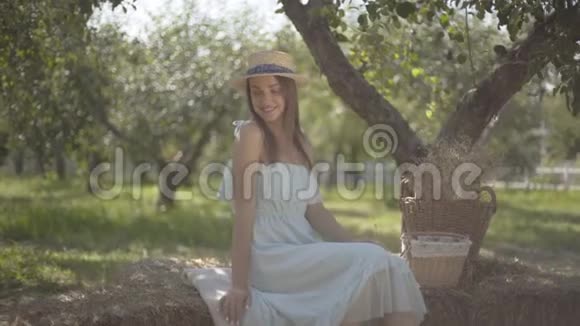 可爱的年轻女子戴着草帽穿着长长的白色连衣裙看着镜头微笑地坐在绿色的夏日花园里视频的预览图