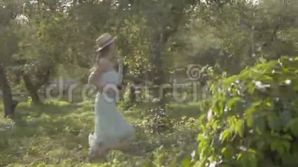 可爱的年轻女子戴着草帽穿着白色的长裙穿过绿色的夏日花园免费农村生活视频的预览图