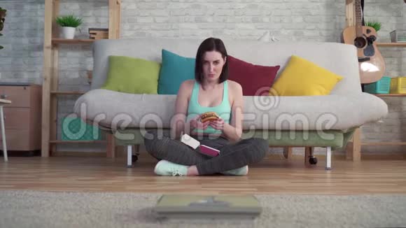 年轻运动的女人坐在地板上看着浴室的鳞片吃汉堡视频的预览图