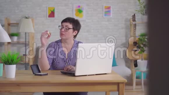 戴眼镜的微笑女人坐在客厅的桌子旁手里拿着助听器她用笔记本电脑视频的预览图