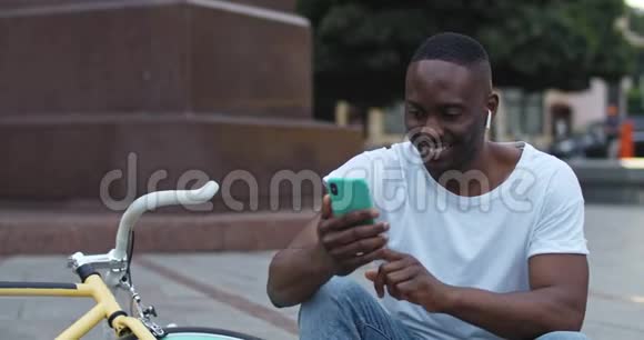令人兴奋的非裔美国人戴着耳机用他的智能手机听音乐边唱边笑视频的预览图